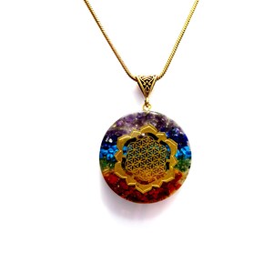 Könnte beinhalten: Eine goldene Kettenkette mit einem runden Anhänger, der mit einem Regenbogen aus Edelsteinen und einem goldenen Lebensblume-Design verziert ist.
