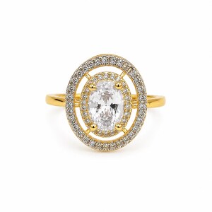 Könnte beinhalten: Ein goldener Ring mit einem zentralen, oval geschliffenen, klaren Stein, der von einem Kranz aus kleinen Pavé-gefassten Steinen umgeben ist. Ein äußerer ovaler Rahmen, ebenfalls mit kleinen Pavé-Steinen besetzt, umschließt den inneren Kranz.