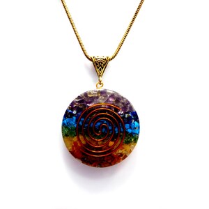 Könnte beinhalten: Eine goldene Kettenkette mit einem runden Anhänger, der eine Kupferspirale umgibt, die von sieben Chakra-Farbensteinen umgeben ist.