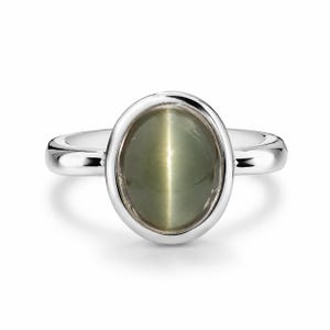 Könnte beinhalten: Ein Ring aus poliertem Silber mit einem glatten, ovalen Cabochon-Edelstein in einem gedämpften Salbeigrün. Der Stein zeigt einen ausgeprägten chatoyierenden Lichtreflex, der ein vertikales Lichtband in der Mitte erzeugt, gefasst in einer Lünette.