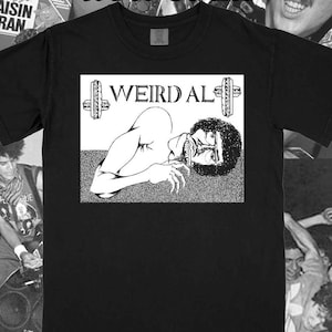 May include: A black t-shirt featuring a white graphic print of a person with curly hair lying down. The text WEIRD AL is printed above the figure, flanked by two vertical sandwich illustrations. The design is set against a textured background.