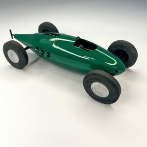 May include: A vintage green toy race car with black tires and silver rims. The car has a streamlined design with a single seat and a small black steering wheel. The toy car is a classic collectible.