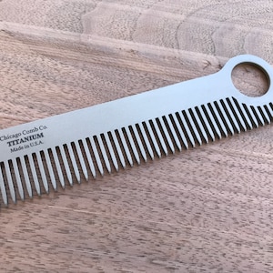 Puede incluir: Un peine de titanio plateado con un agujero en el mango. El peine está grabado con el texto "Chicago Comb Co. TITANIUM Made in U.S.A."
