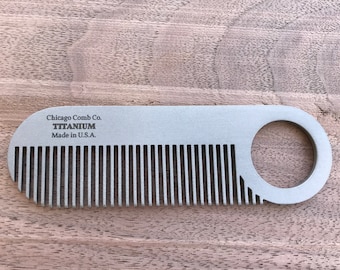 Puede incluir: Un peine de titanio plateado con un asa en forma de bucle. El peine tiene dientes finos y el texto "Chicago Comb Co. TITANIUM Made in U.S.A." está impreso.