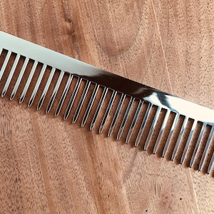 May include: A silver-colored comb with closely spaced teeth. The comb has a rectangular shape with rounded corners and the text "C*" is visible on the top right corner. The comb is lying on a wooden surface.