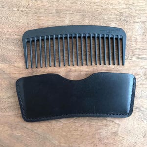 May include: A black comb with fine teeth and a black leather case, set against a wooden surface. The comb is made of carbon fiber and is designed for grooming hair. The case is stitched around the edges.