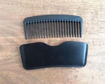 Puede incluir: Un peine negro con dientes finos y un estuche de cuero negro, sobre una superficie de madera. El peine está hecho de fibra de carbono y está diseñado para peinar el cabello. El estuche está cosido en los bordes.