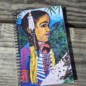 May include: Spiral-bound notebook featuring a colorful illustration of a person with Native American-inspired attire. The artwork includes a profile portrait with a feather, braids, and a horse emblem. The background shows a mountain landscape.