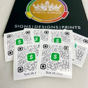 May include: A black mousepad with the text "VINYL KINGS" and the words "SIGNS | DESIGNS | PRINTS" in white. Several white stickers with QR codes and green dollar sign logos are in front of the mousepad. The stickers have the text "$Let_VK_Print".