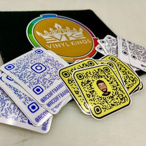 Op de afbeelding: Een stapel witte vierkante stickers met blauwe en zwarte QR-codes. De stickers hebben de tekst "@RCQIFFBARBET" erop gedrukt. Er is ook een gele vierkante sticker met een zwart-witte QR-code en een cartoonafbeelding van een persoon met een baard.