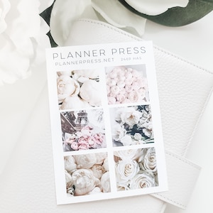 May include: A white card with floral images and the text "PLANNER PRESS PLANNERPRESS.NET 2469 HAS" is displayed on a white leather surface. The card features images of white and pink flowers, including roses and peonies, and the Eiffel Tower.