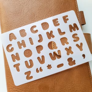 May include: A white plastic alphabet stencil with rounded letters and symbols rests on a brown leather notebook. The stencil includes the letters A through Z, along with punctuation marks. The notebook has a textured surface and a strap.