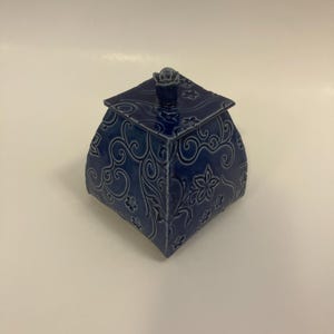 May include: A square, deep blue ceramic jar with a matching lid. The exterior features a raised, embossed floral and swirling vine pattern across all four sides and the lid. A small, sculpted rose-shaped knob sits atop the center of the lid.