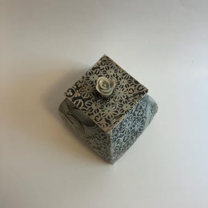 May include: A square ceramic box with a lid featuring a dark gray and white floral pattern. The lid is topped with a small, sculpted three-dimensional ceramic rose in a light gray tone. The box has a textured surface with intricate botanical details.