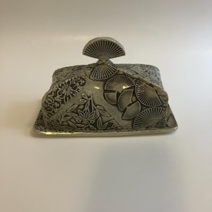 Op de afbeelding: Een rechthoekige keramische botervloot met een bijpassend deksel voorzien van een waaiervormige handgreep. Het oppervlak is versierd met een donkergrijs en wit reliëfpatroon van gestileerde waaiers en bloemmotieven op het deksel en de bodemplaat.