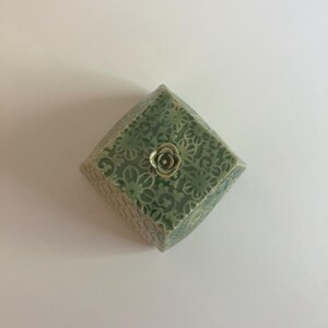 Könnte beinhalten: Eine quadratische Schmuckschatulle aus Keramik mit einem grünen Blumenmuster und einer kleinen, goldfarbenen Rose auf dem Deckel. Die Box hat eine geometrische Form und eine strukturierte Oberfläche, was auf einen handgefertigten Artikel hindeutet.
