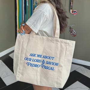 May include: Beige tote bag with blue text that reads "ASK ME ABOUT OUR LORD & SAVIOR PEDRO PASCAL." The bag is carried by a person wearing a pink baseball cap and a white t-shirt. The background features colorful artwork.