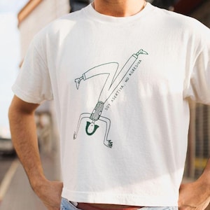 Könnte beinhalten: Weißes, bauchfreies T-Shirt mit einer grünen Illustration einer Person, die einen Handstand macht. Der Text "SOY ASERTIVA, NO AGRESSIVA" steht auf dem Shirt.