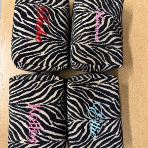 May include: Four rectangular zippered pouches featuring a black and white zebra print pattern. Each pouch is personalized with embroidered names in different colors: red for Sasha, pink for Mushreena, pink for Kelley, and light blue for Ellie.