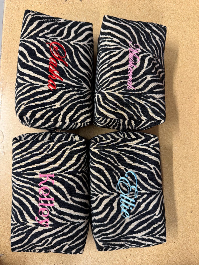 May include: Four rectangular zippered pouches featuring a black and white zebra print pattern. Each pouch is personalized with embroidered names in different colors: red for Sasha, pink for Mushreena, pink for Kelley, and light blue for Ellie.