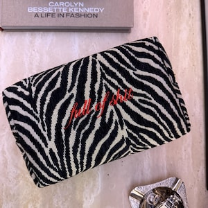 May include: A rectangular zippered pouch featuring a black and white zebra print pattern. The text "full of shit" is embroidered in red cursive script across the center of the pouch. The fabric has a textured, woven appearance.
