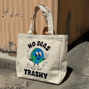 May include: A white canvas tote bag with a cartoon earth wearing cowboy boots and peace signs. The text "No Seas Trashy" is printed on the bag.