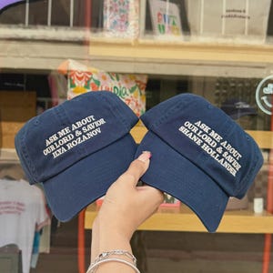 May include: Two navy blue baseball caps with white embroidered text. One reads "ASK ME ABOUT OUR LORD & SAVIOR ILYA ROZANOV", the other "ASK ME ABOUT OUR LORD & SAVIOR SHANE HOLLANDER". The caps are displayed in front of a store window.
