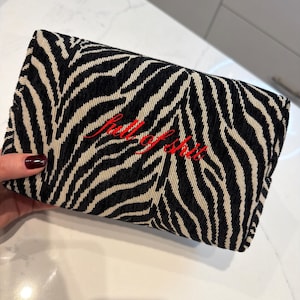 May include: A rectangular clutch bag featuring a black and white zebra stripe pattern. The front of the bag is embroidered with the text "full of shit" in a cursive red font. The fabric has a textured appearance with a structured rectangular shape.