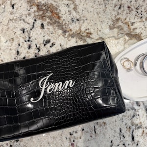 Puede incluir: Bolsa de cosméticos negra de piel sintética con textura de cocodrilo. El nombre "Jenn" se muestra en letras blancas cursivas en la parte delantera. La bolsa es rectangular y parece estar cerrada. Una bandeja blanca con joyas está en el fondo.