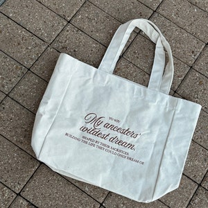 May include: A white canvas tote bag with brown text that reads "YO SOY My ancestors' wildest dream. SHAPED BY THEIR SACRIFICES. BUILDING THE LIFE THEY COULD ONLY DREAM OF."