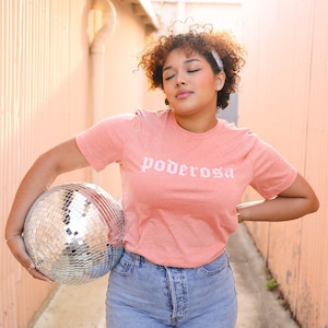 May include: A peach-colored t-shirt with the word "poderosa" in white script. The person is holding a disco ball. The person is wearing blue jeans and a white headband. The background is a peach-colored alley.
