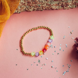 May include: A gold beaded bracelet with colorful beads spelling out the word "PODEROSA". The bracelet is on a pink surface with silver glitter.