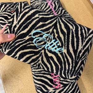 May include: A rectangular fabric pouch featuring a black and beige zebra stripe pattern. The front of the pouch is embroidered with the name Ellie in light blue cursive script. Additional similar pouches with different names are visible nearby.