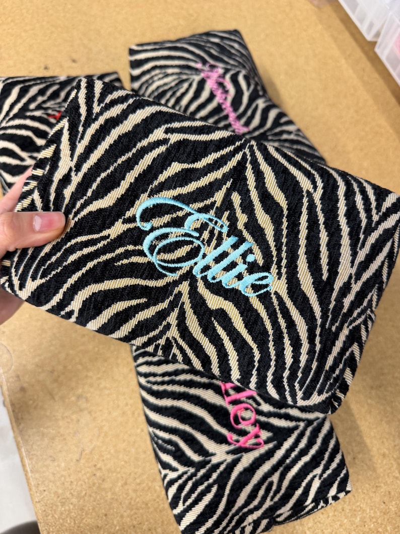 May include: A rectangular fabric pouch featuring a black and beige zebra stripe pattern. The front of the pouch is embroidered with the name Ellie in light blue cursive script. Additional similar pouches with different names are visible nearby.