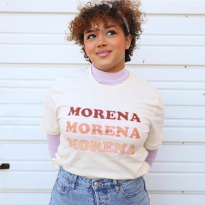 May include: A cream-coloured t-shirt displaying the word "MORENA" in a gradient of brown and peach hues. The t-shirt is worn with light blue denim jeans. The person is also wearing a lilac turtleneck.