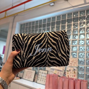May include: A rectangular zippered pouch featuring a black and beige zebra stripe pattern. The front of the pouch is embroidered with the name Jenn in a white cursive script font. The fabric has a textured appearance with a structured rectangular shape.