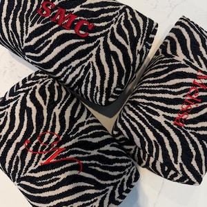 May include: Three rectangular fabric pouches featuring a black and white zebra stripe pattern. Each pouch is embroidered with red thread, displaying the text SMC, the name Melissa, and a cursive letter M respectively on the textured fabric surface.