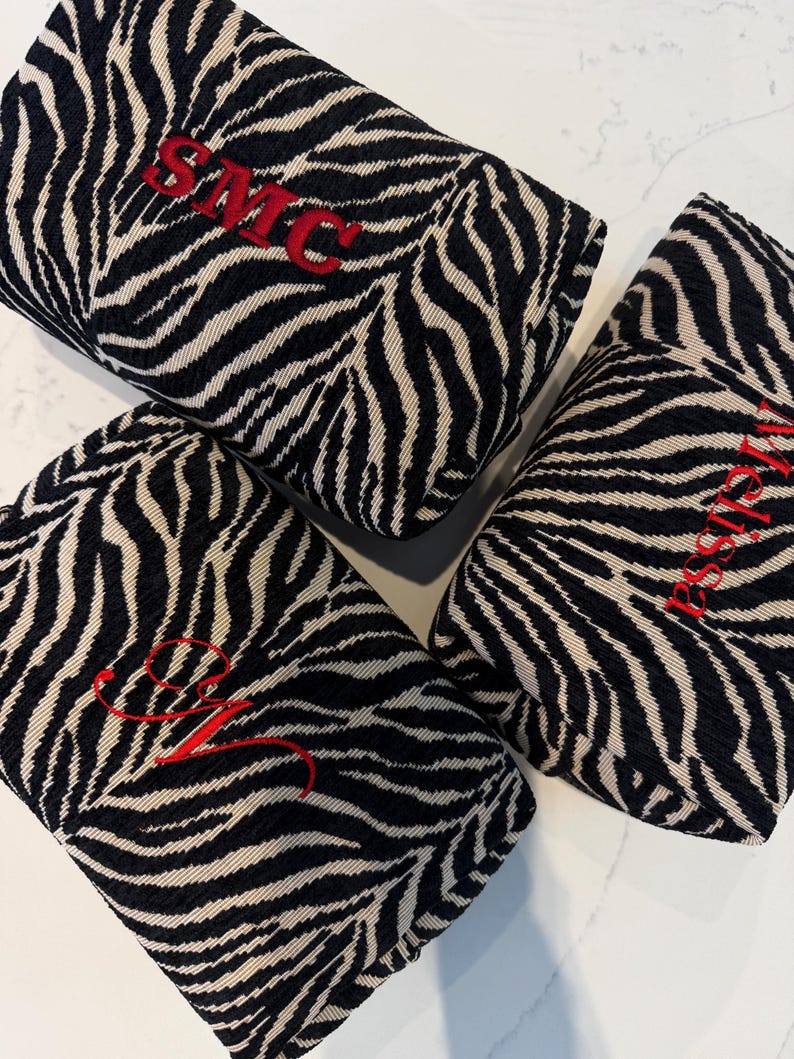 May include: Three rectangular fabric pouches featuring a black and white zebra stripe pattern. Each pouch is embroidered with red thread, displaying the text SMC, the name Melissa, and a cursive letter M respectively on the textured fabric surface.