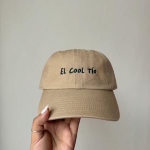 Puede incluir: Gorra de béisbol beige con el texto "EL COOL TÍO" bordado en verde. La gorra tiene una visera curva y un diseño clásico de seis paneles, adecuada para uso casual.