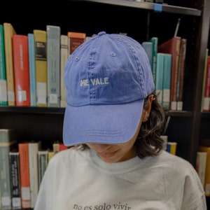 May include: A blue baseball cap with the embroidered text "ME VALE" in white. The cap is worn by a person in front of a bookshelf filled with books. The person is wearing a white t-shirt.