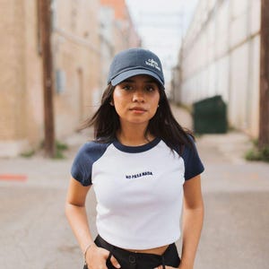 May include: A woman wearing a white and navy blue baseball tee with the text "NO PASA NADA" on the front. She is wearing a dark blue baseball cap with a white logo and black pants.