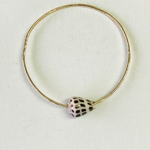 Może przedstawiać: Złota bransoletka typu bangle z małym, białym i brązowym urokiem w kształcie muszli.