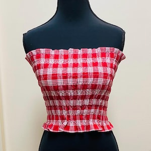 Puede incluir: Top de tubo a cuadros rojos y blancos con dobladillo con volantes. El top se muestra en un maniquí negro. La tela tiene un diseño fruncido y elástico.
