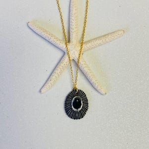 Könnte beinhalten: Eine goldfarbene Halskette mit einem schwarzen und silbernen Anhänger, der einen schwarzen ovalen Stein aufweist. Der Anhänger hat ein Sonnenstrahl-Design. Die Halskette wird auf einem weißen Seestern vor weißem Hintergrund präsentiert.