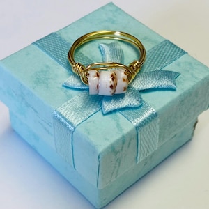 May include: A gold-coloured wire ring featuring three white and brown speckled beads. The ring is presented on a light blue gift box with a matching ribbon and bow. The ring is a piece of jewellery.