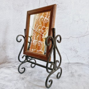 May include: A wooden framed mirror with a floral pattern reflected in the glass, mounted on a black metal stand with ornate scrollwork.