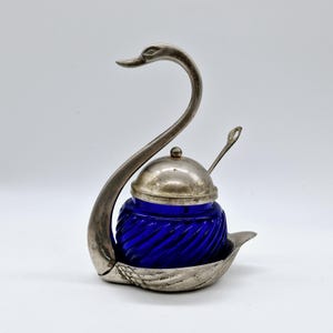 May include: A silver swan-shaped sugar dispenser with a blue glass jar and a lid. The jar has a swirl pattern and the swan has a spoon.