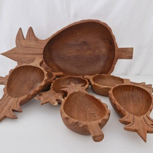 Può includere: Set di sei ciotole di legno a forma di ananas. Le ciotole sono realizzate in legno marrone scuro e hanno un design a forma di ananas intagliato. La ciotola più grande è di forma ovale e le ciotole più piccole sono rotonde.