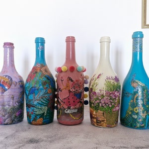 5 Handpainted & decoupaged wine bottles with glitter accents.  
Pink bottle has picture of a girl on a bicycle, and colorful pompons.
Whitish bottle has Provencal floral pattern.
Turquoise bottles, one  has exotic  flower pattern. Second has  peacock