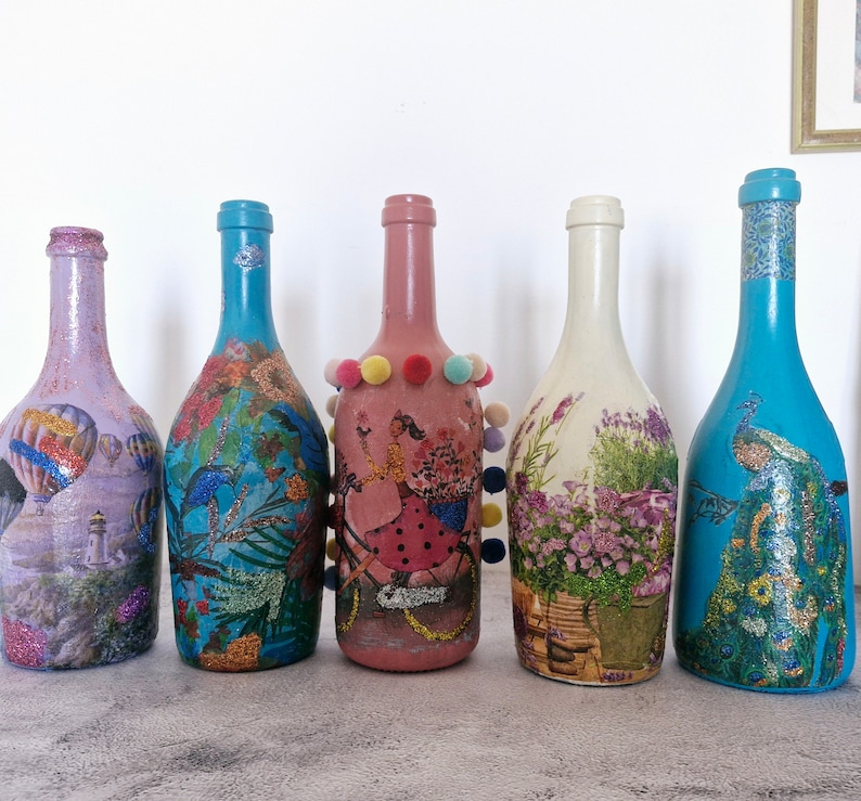 5 Handpainted & decoupaged wine bottles with glitter accents.  
Pink bottle has picture of a girl on a bicycle, and colorful pompons.
Whitish bottle has Provencal floral pattern.
Turquoise bottles, one  has exotic  flower pattern. Second has  peacock